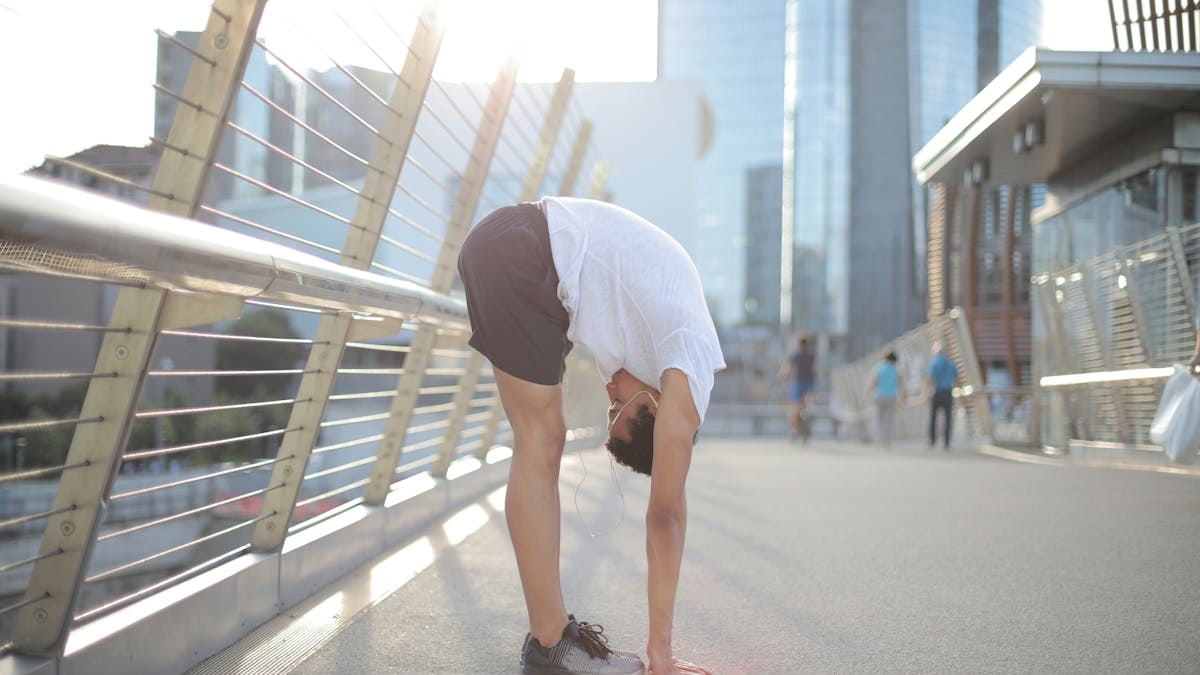 Side view of young African American man in earphones and sportswear listening to music and stretching body while performing standing forward bend exercise on street against blurred high rise buildings in downtown
