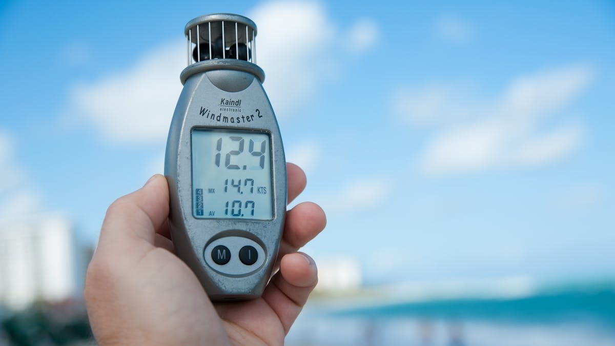Close-up of a hand holding Windmaster 2 anemometer against a clear blue sky and beach backdrop.